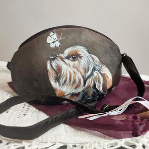May include: A brown fabric handbag with a painted image of a dog looking up at a white butterfly. The bag has a brown strap.