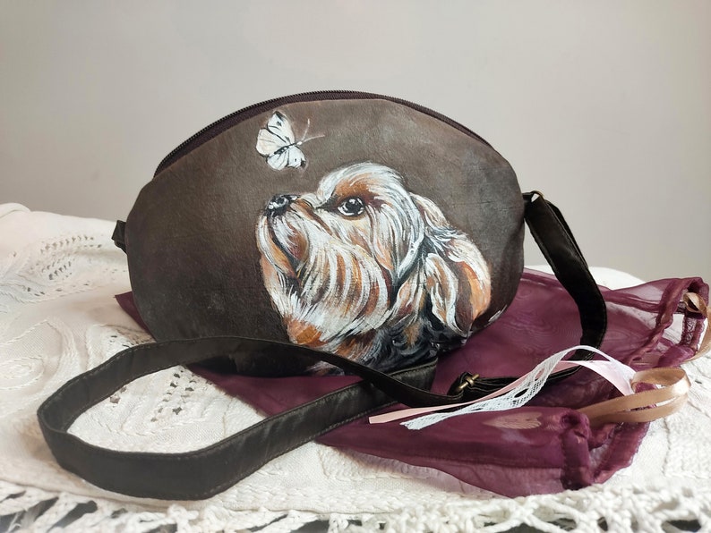 May include: A brown fabric handbag with a painted image of a dog looking up at a white butterfly. The bag has a brown strap.