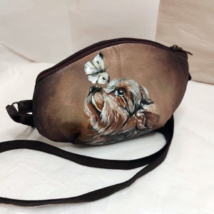 May include: A brown crossbody bag with a painted image of a Yorkshire Terrier looking up at a white butterfly.