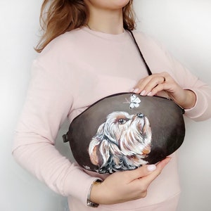 May include: A brown leather crossbody bag with a painted image of a Yorkshire Terrier looking up at a butterfly.