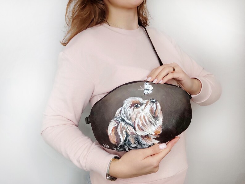 May include: A brown leather crossbody bag with a painted image of a Yorkshire Terrier looking up at a butterfly.