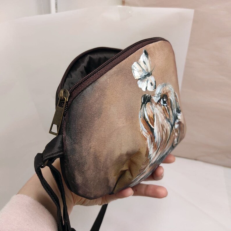 May include: A brown fabric handbag with a zipper closure and a shoulder strap. The bag features a hand-painted design of a dog looking up at a butterfly.
