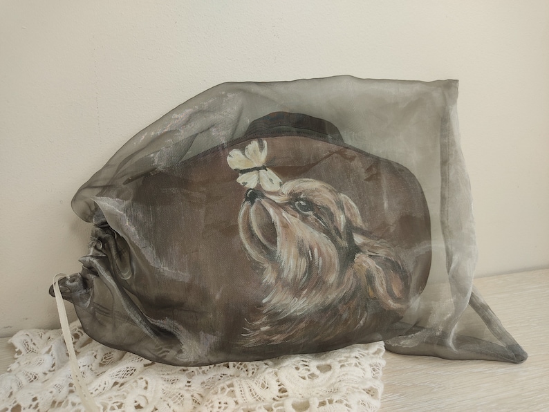 May include: A brown fabric bag with a painted image of a dog looking at a butterfly. The bag is made of a sheer fabric and has a drawstring closure.