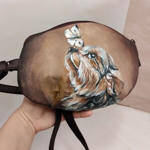 May include: A brown fabric handbag with a painted image of a dog looking up at a butterfly. The dog is brown and black with white markings. The butterfly is white with black markings.