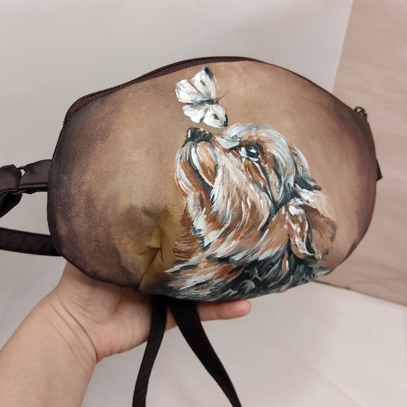 May include: A brown fabric handbag with a painted image of a dog looking up at a butterfly. The dog is brown and black with white markings. The butterfly is white with black markings.