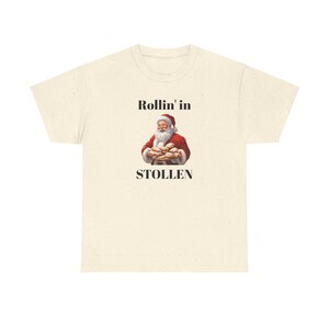 Puede incluir: Una camiseta blanca con un gráfico negro de Santa Claus sosteniendo un plato de pan de especias. El texto "Rollin' in Stollen" está impreso debajo de la imagen.