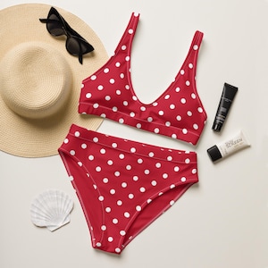 May include: A red bikini set with white polka dots, a straw hat, sunglasses, sunscreen, and a seashell. The bikini top has a V-neck design, and the bottoms are high-waisted.