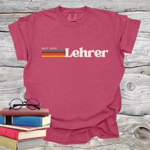 May include: A maroon t-shirt with the word "Lehrer" in white, above a horizontal stripe design in red, orange, yellow, and blue. The text "SEIT 2015" is above the stripes. A stack of books and glasses are in the foreground.