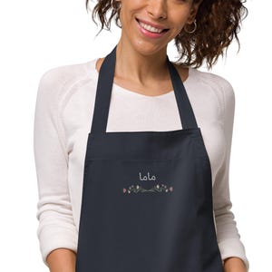 May include: A navy blue apron with the Arabic word "mama" embroidered in white, along with a floral design in pink and green. The apron has a bib and neck strap.