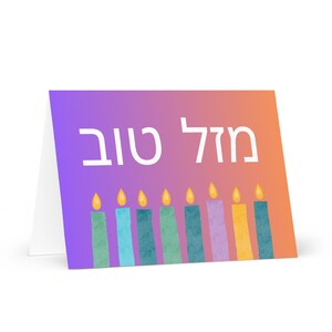 May include: A folded greeting card with a gradient purple and orange background. The card features the Hebrew text "Mazel Tov" in white. Below the text are eight colorful candles with yellow flames, in shades of green, blue, purple, and yellow.
