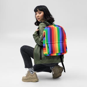 Puede incluir: Una mochila con diseño de rayas verticales de arcoíris. La mochila presenta rayas rojas, naranjas, amarillas, verdes, azules y moradas. Tiene una cremallera y correas negras. Una persona lleva la mochila, vestida con una chaqueta verde y vaqueros negros.