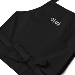 May include: Black apron with white embroidered Korean text. The apron has long ties for securing around the waist and neck. The text reads "아빠".