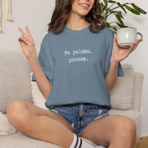 May include: A dusty blue short-sleeved t-shirt featuring the white text "Po polsku, proszę." printed on the center of the chest. The shirt has a crew neckline and a relaxed fit, paired with blue denim shorts.