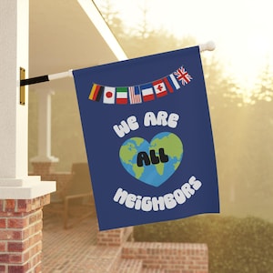 May include: A blue flag with the text "We Are All Neighbors" and a heart-shaped Earth graphic. The flag is decorated with a string of international flags and is mounted on a white pole.