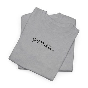 May include: Two light gray t-shirts are neatly folded and stacked. The word "genau" is printed in black, centered on the top shirt. The shirts are made of a soft material and are displayed on a white background.