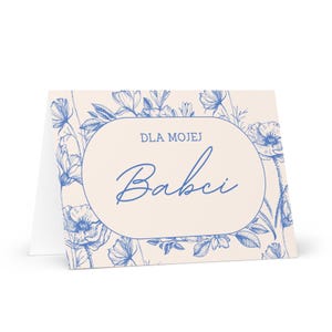 May include: A folded greeting card with a floral design in blue and cream. The card features an oval with the words "DLA MOJEJ Babci" in blue script. The background is cream with blue floral illustrations.