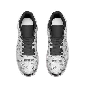 May include: Pair of white high-top sneakers with black laces and a black tongue. The sneakers have a repeating pattern of text and splatters, with the word "RESIST" prominently displayed on the toe.