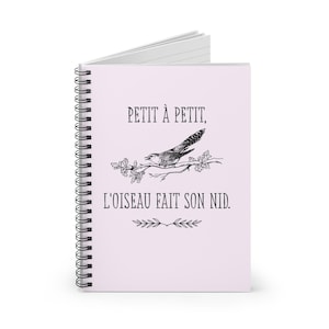 May include: A spiral-bound notebook with a light pink cover. The cover features the text "PETIT À PETIT, L'OISEAU FAIT SON NID." above and below a black and white illustration of a bird on a branch.