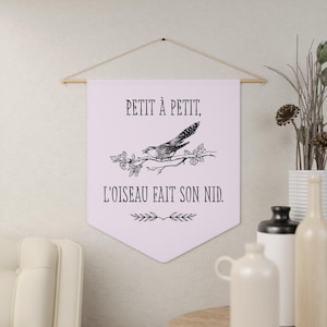 May include: A light purple wall hanging with a bird illustration and the French text "Petit à Petit, L'oiseau Fait Son Nid." The banner is suspended from a wooden rod with a string. Decorative elements include a bird on a branch and floral accents.