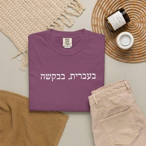 May include: A plum-colored crew neck t-shirt featuring the white Hebrew text "בעברית, בבקשה" printed across the chest. The garment has a visible interior neck label with the brand name Comfort Colors and a size indicator of L-G-G.