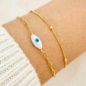 May include: Two gold bracelets on a wrist. One bracelet features a white evil eye charm with a blue centre. The other bracelet is a delicate chain with small gold beads.