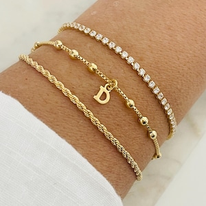 Könnte beinhalten: Drei goldene Armbänder, die an einem Handgelenk übereinander liegen. Das obere Armband ist eine dünne Kette mit kleinen, klaren Steinen. Das mittlere Armband ist eine zarte Kette mit kleinen goldenen Perlen. Das untere Armband ist eine dickere Seilkette mit einem goldenen Anhänger, auf dem "D" steht.
