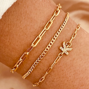 Könnte beinhalten: Drei goldene Kettenarmbänder an einem Handgelenk. Das obere Armband ist eine dicke, klobige Kette. Das mittlere Armband ist eine dünnere, feinere Kette. Das untere Armband hat einen kleinen, mit Diamanten besetzten Buchstabenanhänger.