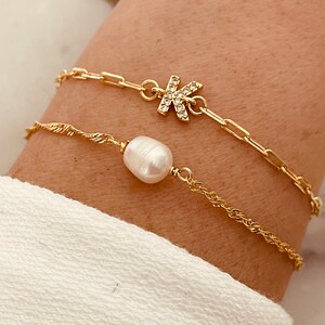 May include: Two gold bracelets on a wrist. One bracelet has a pearl charm and the other has a letter K charm with rhinestones.