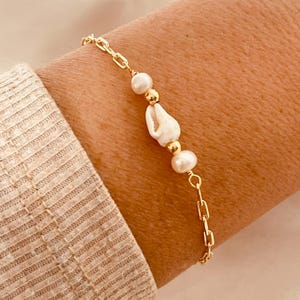 May include: Gold-toned chain bracelet featuring a seashell, pearls, and gold beads. The bracelet is composed of a delicate chain with a lobster clasp. The bracelet is worn on a wrist.