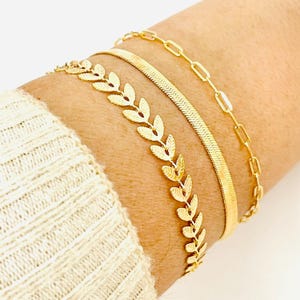 Könnte beinhalten: Drei goldene Armbänder, die an einem Handgelenk übereinander liegen. Das obere Armband ist ein zartes Gliederketten-Design. Das mittlere Armband ist ein dickes, strukturiertes Band. Das untere Armband hat ein sich wiederholendes Blattdesign.