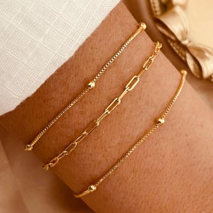 May include: Three gold chain bracelets with small round beads on a person's wrist. The bracelets are layered on top of each other.