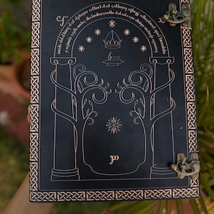 Puede incluir: Diario de cuero negro con un diseño decorativo dorado. La portada presenta un diseño arqueado con estrellas, una corona y texto. El diario tiene un cierre de broche y un borde de nudo celta.