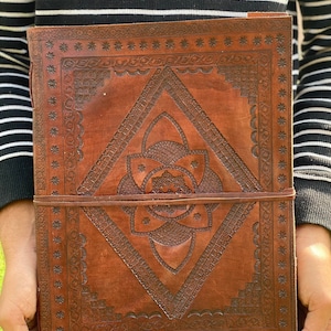 May include: A brown leather journal with intricate embossed designs. The cover features a geometric pattern with a central flower motif. The journal has a leather strap closure.