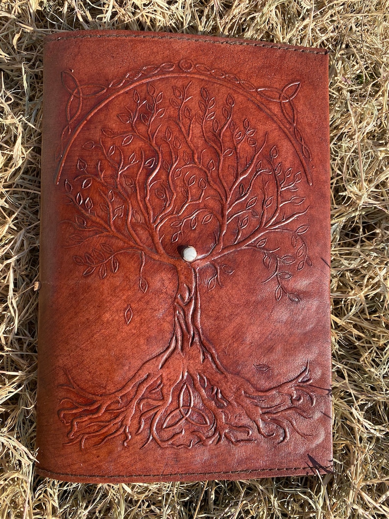 May include: A brown leather journal with a tooled design of a tree of life with intricate roots and branches. The tree is surrounded by a Celtic knot design.