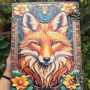 May include: A decorative journal featuring a fox illustration. The cover displays a detailed portrait of a fox in orange, yellow, and white, framed by floral and ornamental designs. The journal has a clasp closure.