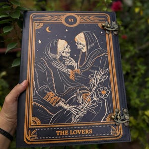 May include: A dark blue tarot card journal with a gold border and clasp. The cover features an illustration of two skeletons in cloaks, with the words "THE LOVERS" at the bottom. The journal is held open, revealing the detailed artwork.