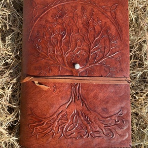 May include: A brown leather journal with a tree of life design embossed on the cover. The tree has intricate branches and roots, and a Celtic knot design is at the top of the cover.