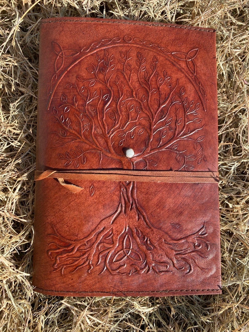 May include: A brown leather journal with a tree of life design embossed on the cover. The tree has intricate branches and roots, and a Celtic knot design is at the top of the cover.