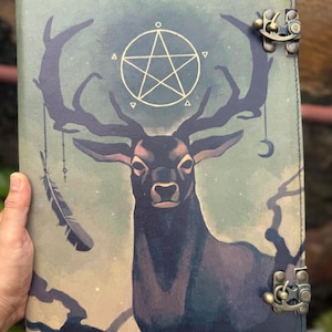 Puede incluir: Un diario de cuero con un diseño místico. La portada presenta un ciervo con grandes cuernos, un pentagrama y amuletos colgantes. La paleta de colores incluye tonos de verde, azul y marrón, con cierres de latón antiguo.