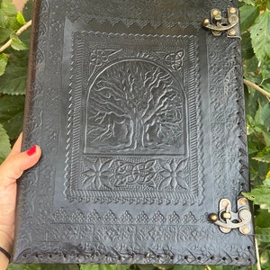 Könnte beinhalten: Schwarzes Lederjournal mit einem geprägten Lebensbaum-Design auf dem vorderen Umschlag. Das Journal hat zwei Metallverschlüsse und dekorative Nähte an den Rändern.