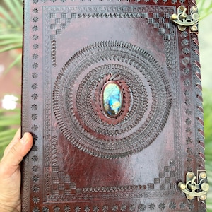 May include: A handcrafted, dark brown leather journal with intricate embossed designs and a central oval gemstone. The journal features a decorative border, metal clasps, and a stitched spine. The gemstone has blue, green, and gold hues.