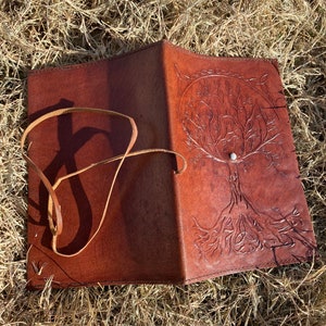 May include: A brown leather journal with a tree of life design embossed on the cover. The journal has a strap closure and a white stone accent.