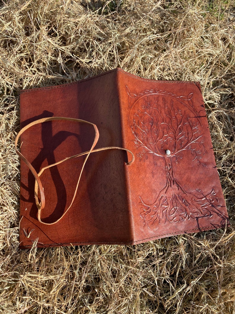May include: A brown leather journal with a tree of life design embossed on the cover. The journal has a strap closure and a white stone accent.