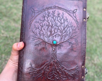 May include: A brown leather journal with a tooled design of a tree of life with a turquoise stone at the center. The journal has a Celtic knot design at the top and is closed with metal clasps.