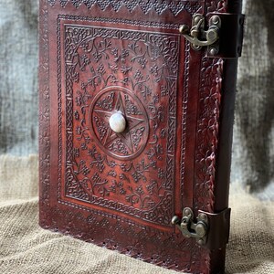 May include: A brown leather journal with intricate floral and Celtic knotwork designs. The cover features a pentacle with a white stone in the center. The journal has two brass clasps.