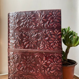 Puede incluir: Un diario de cuero marrón con un patrón floral en relieve. El diario tiene un cierre de correa de cuero marrón.