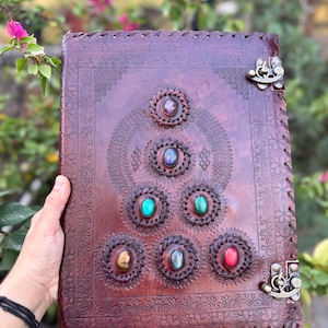 May include: A handcrafted, brown leather journal with decorative stitching and metal clasps. The cover features an embossed design and is adorned with seven colorful gemstones, including red, green, and blue stones, set in circular frames.