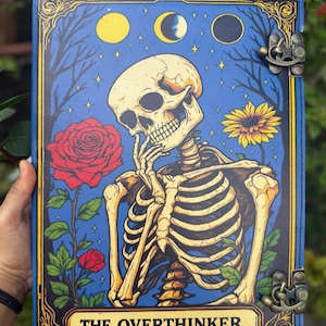 May include: A blue journal with a skeleton illustration, roses, and a sunflower. The cover features the text "THE OVERTHINKER" in a gold frame. The journal has a brass clasp and a decorative border with moon phases.