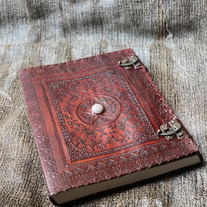 May include: A brown leather journal with a pentagram design and a white stone in the centre. The journal has two metal clasps and is closed.