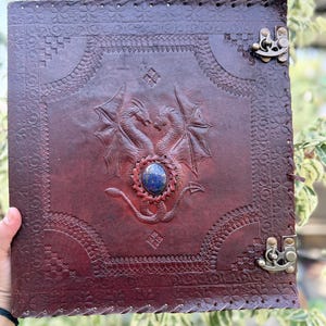 May include: A dark brown leather journal featuring an embossed design of two facing dragons with wings spread. A smooth, oval blue stone is set in the center between the dragons. The cover has decorative border patterns and two metal latches.